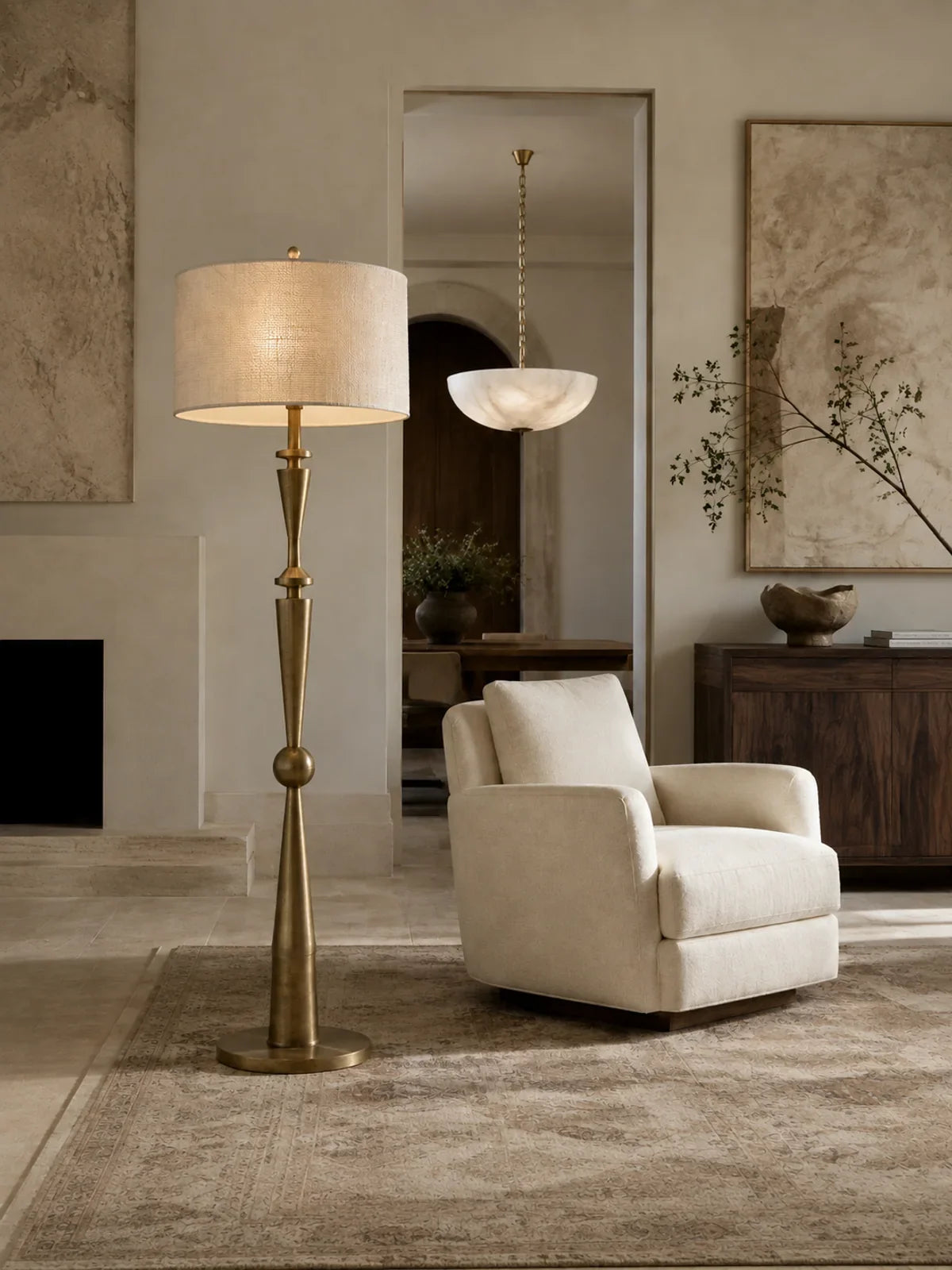 A sculptural aged brass floor lamp with a beautifully woven ivory linen shade in the foreground of a refined living room.