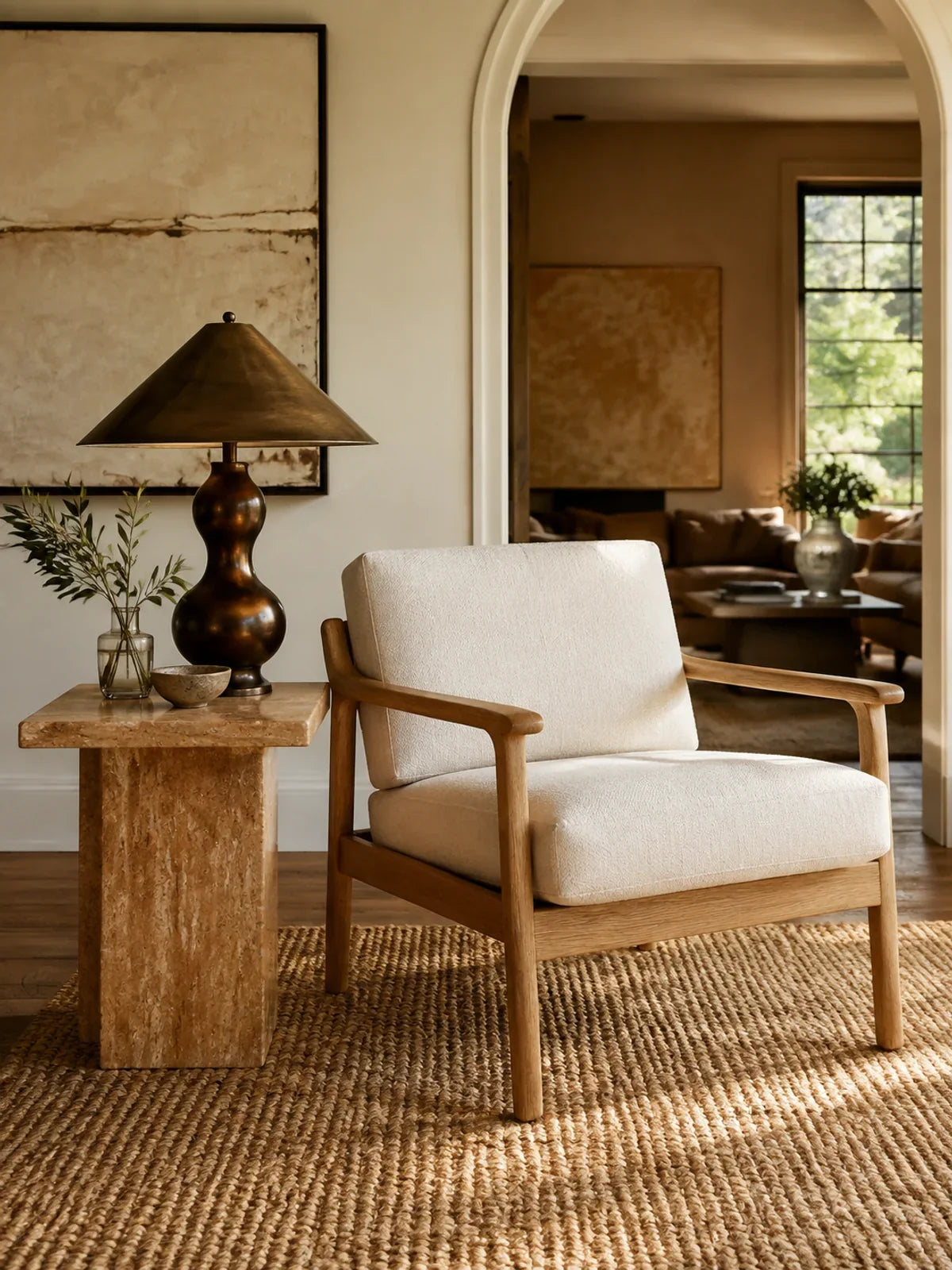 A beautifully curated newly styled living room vignette: one contemporary ivory upholstered oak armchair beside a chunky honey-toned travertine side table, a sculptural bronze lamp and a handwoven rug.