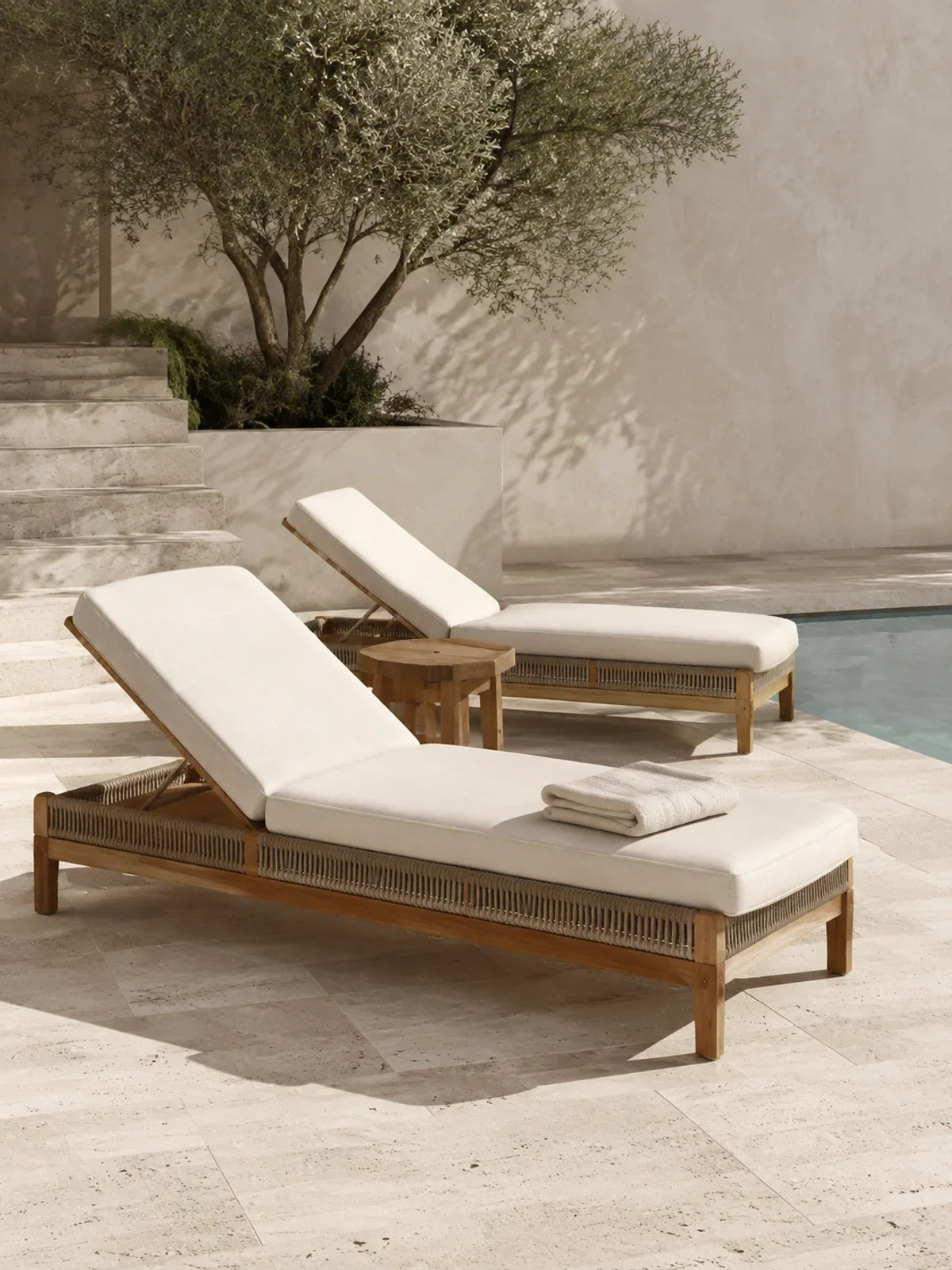 Two premium teak and woven-rope sun loungers with thick off-white cushions beside a still pale-stone pool.
